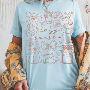 Peut inclure: T-shirt bleu clair avec un motif sur le th&egrave;me de l'automne. Le motif comprend une citrouille, des n&oelig;uds, une tasse de caf&eacute;, une &eacute;charpe, un chapeau et les mots "cozy season". Le mot "Chambray" est en bas.