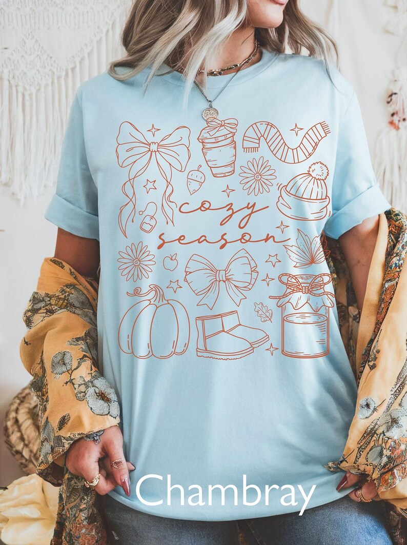 Peut inclure: T-shirt bleu clair avec un motif sur le th&egrave;me de l'automne. Le motif comprend une citrouille, des n&oelig;uds, une tasse de caf&eacute;, une &eacute;charpe, un chapeau et les mots "cozy season". Le mot "Chambray" est en bas.
