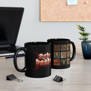 Puede incluir: Dos tazas de cerámica negras. Una taza presenta el texto "Don't Panic" en una fuente roja y naranja con un diseño de rayo. La otra taza tiene una cuadrícula de cuadrados ilustrados con varias imágenes, incluyendo armas, un rayo y una calavera.