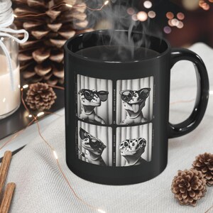 Könnte beinhalten: Schwarze Keramik-Tasse mit einem Vier-Panel-Design einer Eidechse mit Sonnenbrille. Die Eidechsenbilder sind in Schwarzweiß gehalten. Die Tasse hat einen gebogenen Griff und ist mit einem dampfenden Getränk gefüllt. Dekorative Tannenzapfen und eine Kerze sind im Hintergrund.
