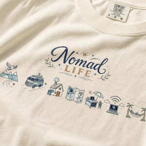 May include: Cream-colored t-shirt with the words "Nomad Life" in navy blue script. The design includes illustrations of a van, mountains, a house, a map, a laptop, and a hammock.