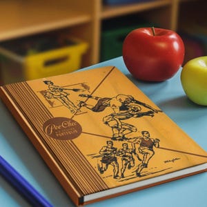 May include: A vintage "Pee-Chee" portfolio notebook with illustrations of athletes in action. The cover is tan with brown and black drawings. A red apple and a green apple sit nearby on a blue surface. A blue pen is in the foreground.