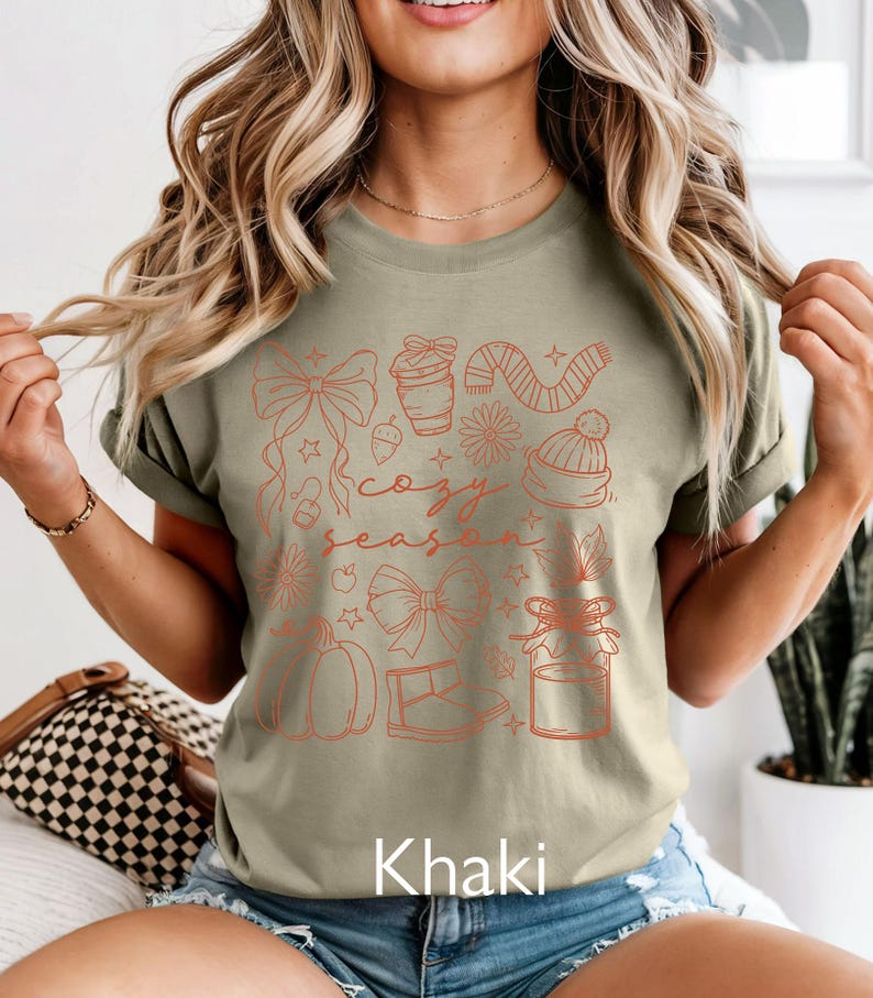 Peut inclure: T-shirt kaki avec un motif imprim&eacute; couleur rouille. Le motif comprend des n&oelig;uds, une tasse &agrave; caf&eacute;, une &eacute;charpe, un bonnet, une citrouille et l'inscription "cozy season".