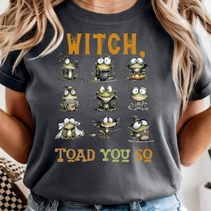 May include: Dark grey t-shirt with the words "WITCH, TOAD YOU SO" in orange and green. The shirt features cartoon frogs in Halloween costumes. The frogs are in various poses, some with witch hats and capes.