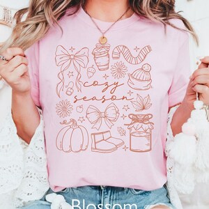 Peut inclure: T-shirt rose clair avec un motif graphique marron repr&eacute;sentant des illustrations sur le th&egrave;me de l'automne, dont une citrouille, un n&oelig;ud et un pot de confiture. Les mots "cozy season" sont &eacute;crits en cursive. Le mot "Blossom" est imprim&eacute; en bas.