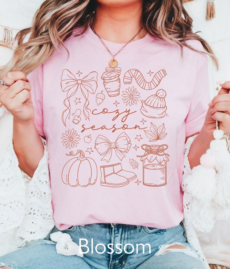 Peut inclure: T-shirt rose clair avec un motif graphique marron repr&eacute;sentant des illustrations sur le th&egrave;me de l'automne, dont une citrouille, un n&oelig;ud et un pot de confiture. Les mots "cozy season" sont &eacute;crits en cursive. Le mot "Blossom" est imprim&eacute; en bas.