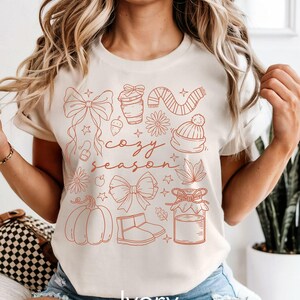 Peut inclure: T-shirt ivoire avec un motif rouille repr&eacute;sentant des illustrations sur le th&egrave;me de l'automne. Le motif comprend une citrouille, un n&oelig;ud, une tasse &agrave; caf&eacute;, une &eacute;charpe, un chapeau et les mots "cozy season".