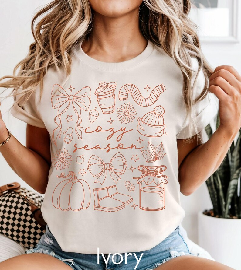 Peut inclure: T-shirt ivoire avec un motif rouille repr&eacute;sentant des illustrations sur le th&egrave;me de l'automne. Le motif comprend une citrouille, un n&oelig;ud, une tasse &agrave; caf&eacute;, une &eacute;charpe, un chapeau et les mots "cozy season".