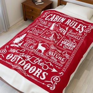 May include: A red and white cabin-themed blanket with the words "Cabin Rules" and other phrases like "Enjoy the Outdoors" and "Take a Long Nap." The blanket features illustrations of trees, a deer, and a campfire.