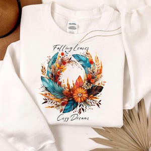 May include: A white long-sleeve sweatshirt featuring a central graphic of a wreath made of colorful feathers, autumn leaves, and a pumpkin. The text "Falling Leaves" is printed above the wreath, and "Cosy Dreams" is printed below the graphic.