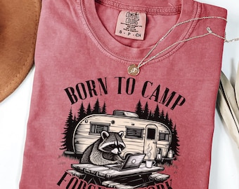 May include: A dusty rose-colored t-shirt with the words "BORN TO CAMP FORCE TO WORK" printed in black. The graphic features a raccoon working on a laptop at a picnic table in front of a camper. A silver necklace with a pendant is draped across the shirt.