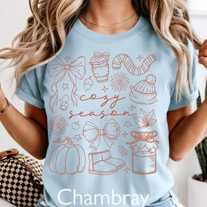 Peut inclure: T-shirt bleu clair avec un motif sur le th&egrave;me de l'automne de couleur rouille. Le motif comprend une citrouille, des bottes, une &eacute;charpe et les mots "cozy season". Le mot "Chambray" est imprim&eacute; en bas.
