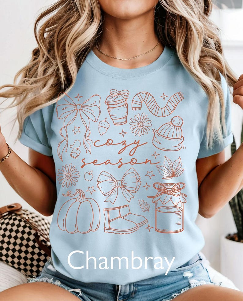 Peut inclure: T-shirt bleu clair avec un motif sur le th&egrave;me de l'automne de couleur rouille. Le motif comprend une citrouille, des bottes, une &eacute;charpe et les mots "cozy season". Le mot "Chambray" est imprim&eacute; en bas.