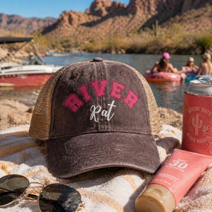 May include: A brown baseball cap with the text "River Rat" in pink lettering. The cap has a mesh back and is on a towel with sunglasses and sunscreen. A can of "Desert Days River Nights" is also visible.