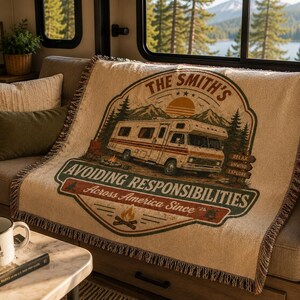 May include: A woven throw blanket featuring a graphic of a recreational vehicle, mountains, and a campfire. The text reads THE SMITH'S, AVOIDING RESPONSIBILITIES, Across America Since '26, RELAX, UNWIND, EXPLORE. The blanket has a fringed edge.