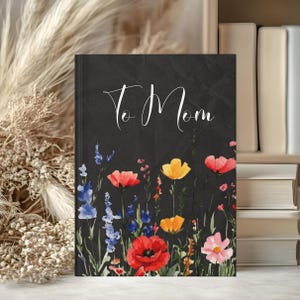 May include: A black journal with the words "To Mom" in white script. The bottom of the journal features a watercolor floral design with red poppies, yellow and orange flowers, and blue wildflowers.