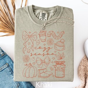 Peut inclure: Sweat-shirt kaki avec un motif "Cozy Season" orange. Le motif comprend des illustrations de citrouille, n&oelig;ud, tasse &agrave; caf&eacute;, &eacute;charpe et autres &eacute;l&eacute;ments d'automne. Un collier argent&eacute; est pos&eacute; sur le sweat-shirt.
