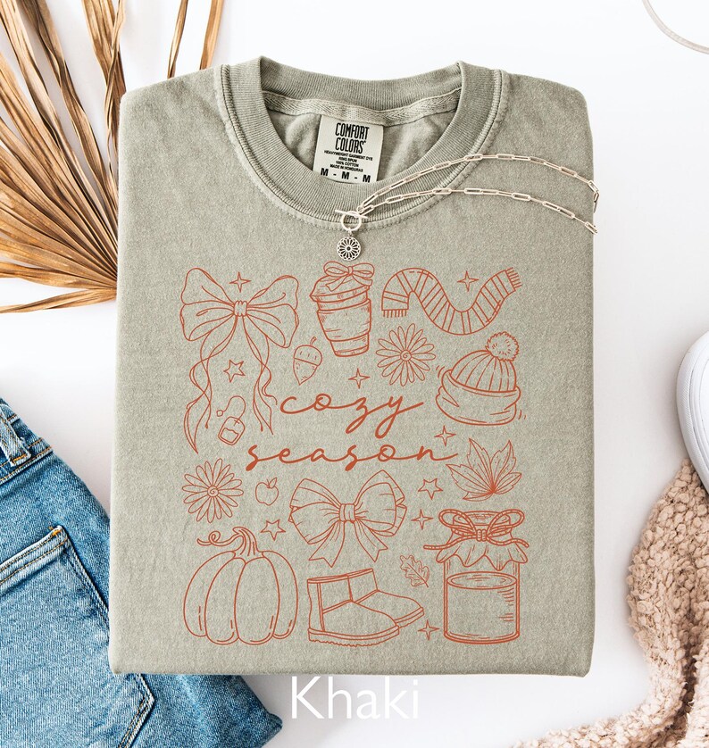 Peut inclure: Sweat-shirt kaki avec un motif "Cozy Season" orange. Le motif comprend des illustrations de citrouille, n&oelig;ud, tasse &agrave; caf&eacute;, &eacute;charpe et autres &eacute;l&eacute;ments d'automne. Un collier argent&eacute; est pos&eacute; sur le sweat-shirt.