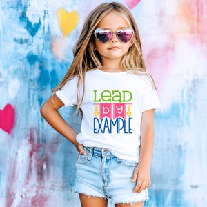 May include: A white t-shirt with a green, pink, and blue graphic that says "Lead by Example". The shirt is being worn by a young person with long blonde hair and heart-shaped sunglasses.