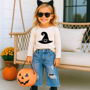 May include: A long-sleeved cream-coloured shirt featuring a black graphic of a witch hat, paired with distressed blue denim jeans, black cat ear headband, black sunglasses, and an orange plastic jack-o'-lantern bucket for trick-or-treating.