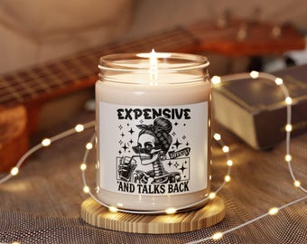 May include: A clear glass jar candle with a lit wick, featuring a white label with a black graphic of a skeleton wearing sunglasses and a headband, drinking from a glass. The text reads EXPENSIVE AND TALKS BACK with the word DIFFICULT on a banner.