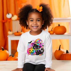 May include: A white long-sleeve shirt featuring a colorful graphic of a black cat wearing a rainbow-patterned witch hat and bow tie, sitting next to a bright pumpkin, surrounded by small stars, candy, and bats on a white fabric background.