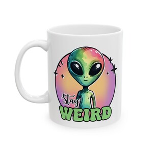 May include: White ceramic mug featuring a colorful illustration of an alien with large black eyes. The alien is set against a pink and yellow background, with the words "Stay Weird" in green and purple. The mug has a white handle.