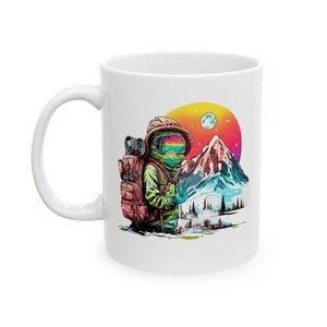 Op de afbeelding: Witte keramische mok met een kleurrijke illustratie van een astronautachtig figuur met een rugzak, bril en berglandschap. Het ontwerp omvat een berg, bomen en een levendige zonsondergang. De mok heeft een gebogen handvat.