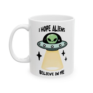 May include: White ceramic mug with a cartoon alien and UFO design. The mug has the text "I HOPE ALIENS BELIEVE IN ME". The design includes a green alien, a light beam, and stars. A fun, quirky design.
