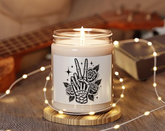 May include: A clear glass jar candle with a white label featuring a black ink illustration of a skeleton hand making a peace sign surrounded by roses and small four-pointed stars. The candle is lit and sits on a round wooden coaster.