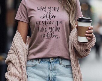 May include: A woman wearing a mauve t-shirt with the text "May your coffee be stronger than your toddler." She is holding a white to-go cup with a brown lid.