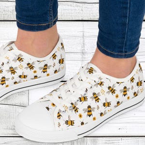 Può includere: Sneakers basse in tela bianca con un motivo di api gialle e nere e fiori bianchi.