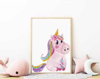 May include: Watercolor print of a pink unicorn with a golden horn, rainbow mane and tail, and floral accents. The framed artwork is displayed on a white wall, with a pink whale, star, and moon-shaped pillow in the foreground.