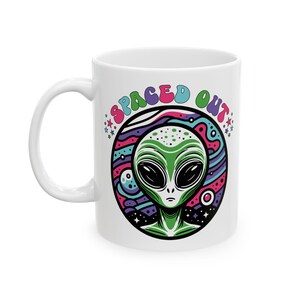 Puede incluir: Taza de cerámica blanca con un diseño de alienígena verde y las palabras "SPACED OUT". El alienígena tiene grandes ojos negros y está sobre un fondo colorido. La taza tiene un asa blanca.