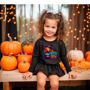 May include: A black long-sleeve dress featuring a graphic print of a cartoon witch with red hair wearing a purple hat, standing next to a purple cauldron with green liquid, and the text "Witch's Brew" in colorful, stylized lettering.