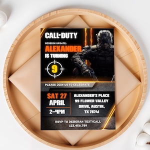 May include: A Call of Duty-themed invitation with the text "Alexander is Turning 9." The invitation features a soldier graphic, orange and yellow accents, and event details. It is set on a wooden tray with an envelope.