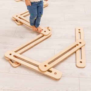 May include: A child stands on a wooden balance beam toy. The toy is made of light-colored wood and has a zig-zag design. The child is wearing a long-sleeved shirt and jeans. The background includes a white wall, a shelf, and decorative items.