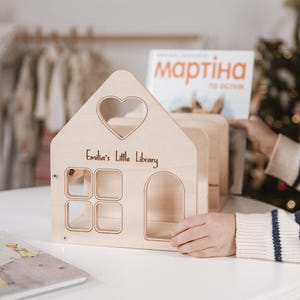 Könnte beinhalten: Ein Bücherregal in Form eines Holzpuppenhauses mit einem herzförmigen Fenster und dem Text "Emilia's Little Library". Das Bücherregal hat vier quadratische Fenster und einen Bogeneingang. Ein Buch liegt auf dem Tisch.