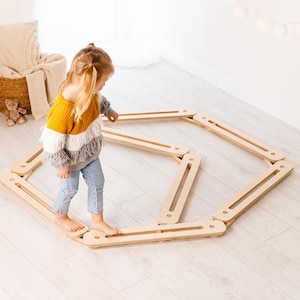 May include: A child in a yellow, white, and grey sweater and blue jeans walks on a wooden balance beam. The balance beam is made of light-coloured wood and is arranged in an octagonal shape on a light-coloured wooden floor. Toys and decor are in the background.