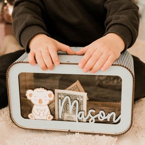 May include: A light blue and wood-toned money box with a clear front panel. Inside, there are dollar bills. A wooden koala and the name "Mason" are on the front. The box is rectangular with rounded edges.