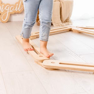May include: A child is walking on a set of wooden balance beams. The beams are light-coloured and rectangular with rounded cutouts. The child is wearing light blue jeans and a grey sweater.