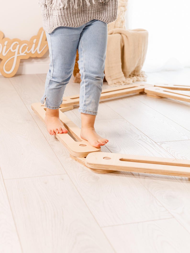 May include: A child is walking on a set of wooden balance beams. The beams are light-coloured and rectangular with rounded cutouts. The child is wearing light blue jeans and a grey sweater.