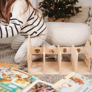 May include: A wooden bookshelf with four sections, decorated with engraved images and cut-out details. Children's books are scattered around the bookshelf on a grey rug. A person wearing a striped sweater is in the background.