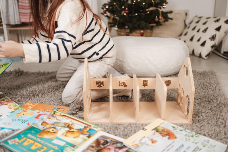 May include: A wooden bookshelf with four sections, decorated with engraved images and cut-out details. Children's books are scattered around the bookshelf on a grey rug. A person wearing a striped sweater is in the background.