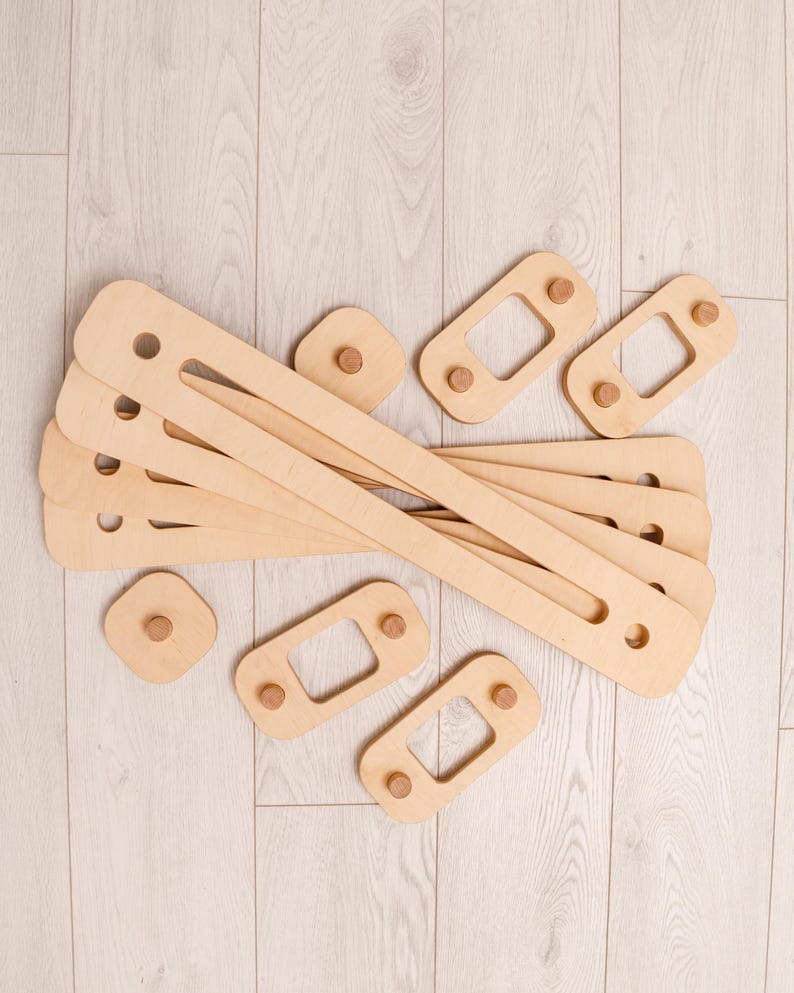 May include: A selection of unfinished wooden toy components. There are long, rectangular pieces with holes, alongside smaller square and rectangular pieces with wooden pegs. The light brown pieces are likely for a construction toy.