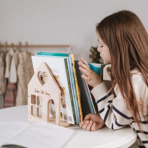 May include: A wooden house-shaped bookend with a heart cutout and the text "Kinder's Little Library." The bookend holds several books. A child wearing a striped sweater is looking at the books. The bookend is on a white table.
