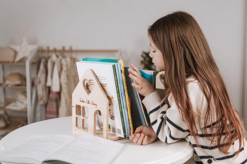 May include: A wooden house-shaped bookend with a heart cutout and the text "Kinder's Little Library." The bookend holds several books. A child wearing a striped sweater is looking at the books. The bookend is on a white table.