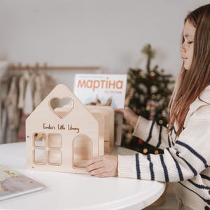 May include: A wooden dollhouse-shaped bookshelf with a heart-shaped window and the text "Emilia's Little Library". The bookshelf has four square windows and an arched doorway. A book is on the table.