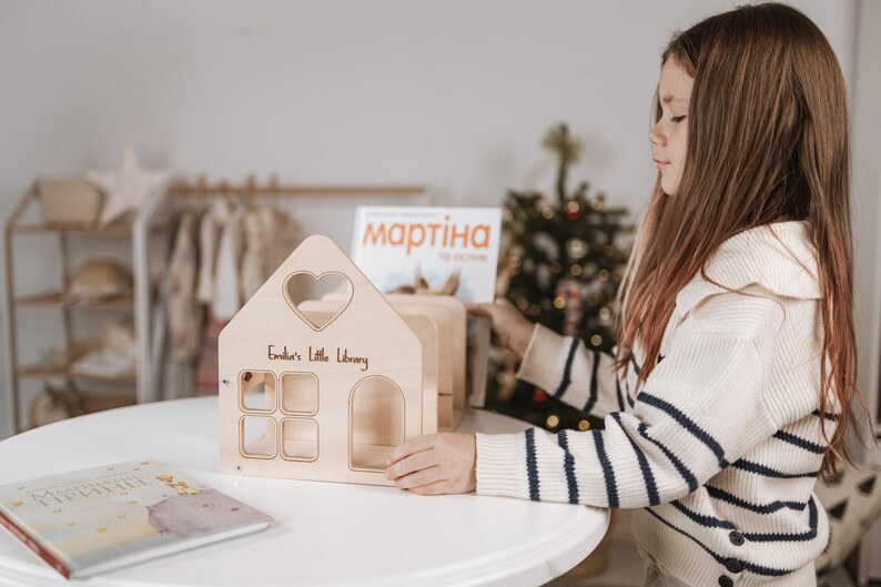 May include: A wooden dollhouse-shaped bookshelf with a heart-shaped window and the text "Emilia's Little Library". The bookshelf has four square windows and an arched doorway. A book is on the table.