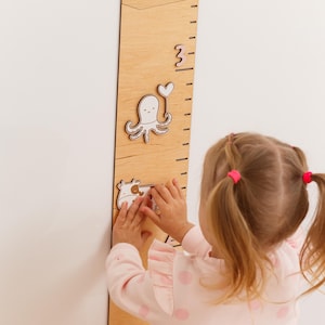 May include: A wooden growth chart with etched height markings and decorative animal cutouts. The chart features an octopus, a heart, and a cow. A young child is interacting with the chart, adding the cow cutout.